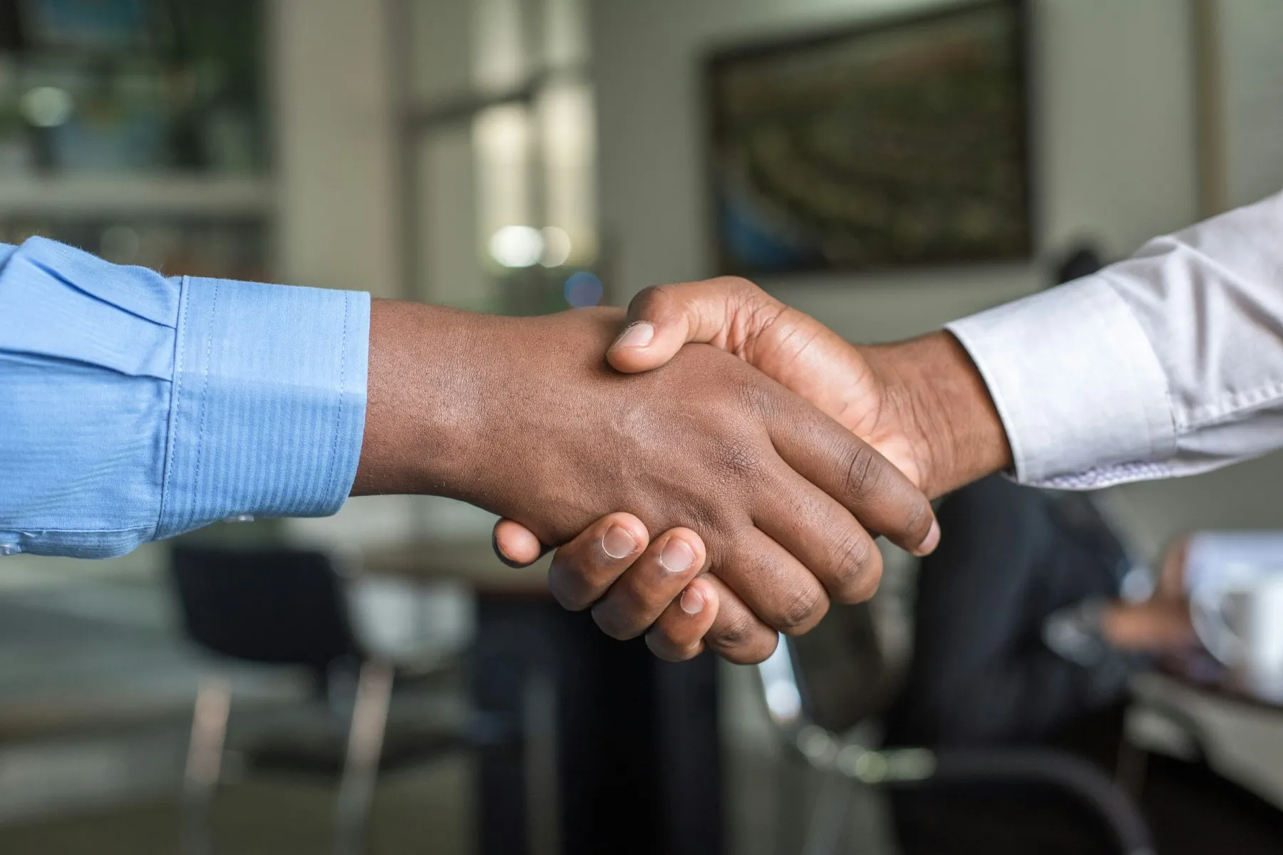 Two people wearing dress shirts shake hands in an office setting, with blurred furniture and a person in the background.