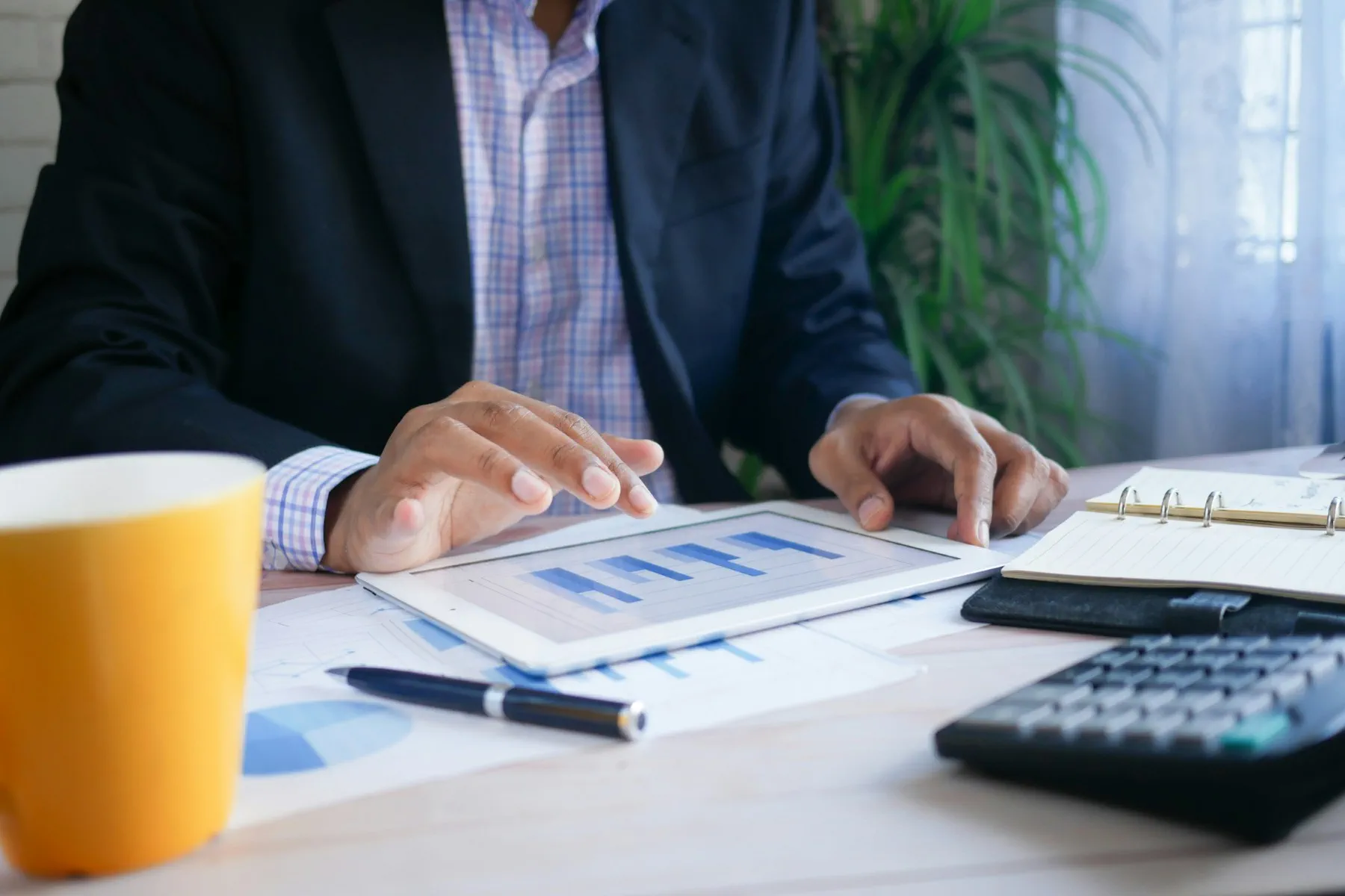 Person in a suit using a tablet to review bar charts at a desk with documents, a pen, a calculator, a notebook, and a yellow mug.