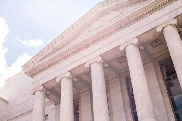The image shows the exterior of a large government or courthouse building with tall marble columns and a triangular pediment under a bright sky.