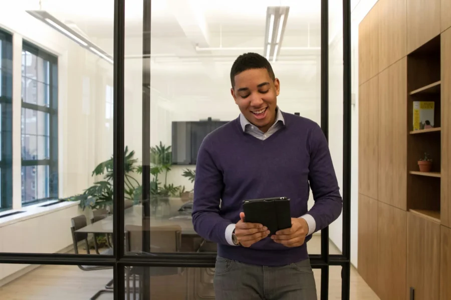 A man standing in an office, smiling while looking at a tablet, with a glass-walled meeting room and plants in the background.