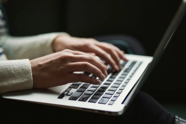 A person types on a laptop keyboard with both hands, wearing a light-colored long-sleeve shirt.